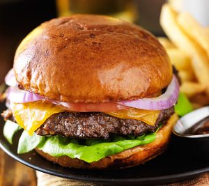 Cheesebuger with lettuce, cheeese, onions, and tomato on a wheat bun alongside blurred out fries in the background.